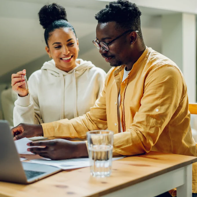 Young man and woman couple smile as they work together at a table with a laptop, papers, and a glass of water.