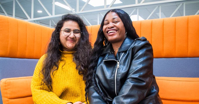 Two students sit next to each other on an orange sofa smiling at the camera.