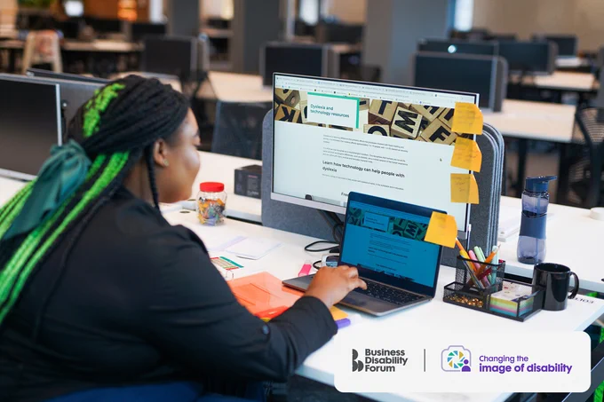 Person browses online dyslexia resources using a laptop and computer screen in an office. There are post-it notes on both screens. The Business Disability Forum 'Changing the image of disability' logo is in the lower right corner of the image.