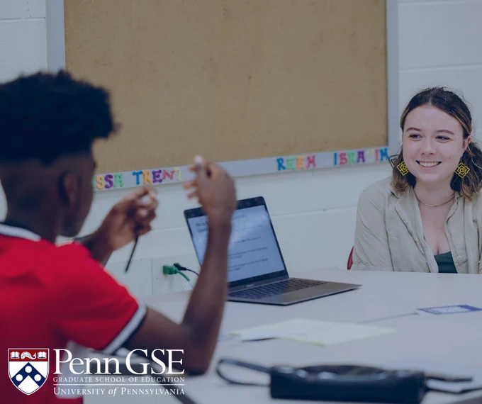 Two students engaged in a discussion at a table with a laptop and books, in a classroom setting at the University of Pennsylvania Graduate School of Education. One student is listening intently while the other gestures during the conversation. The School's logo appears in the corner. 