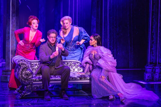Death Becomes Her on Broadway cast with Christopher Sieber sitting, holding a potion, Michelle Williams beside him in purple flowing dress, and Jennifer Simard and Megan Hilty looking upon the potion from behind the chaise. 