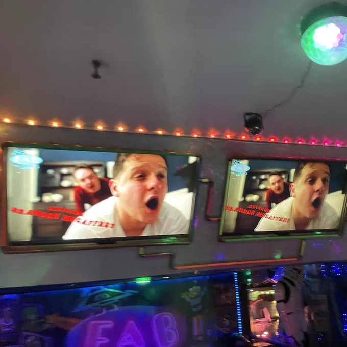 Screenshot of Secrets of a Wallaby Boy trailer – Tim looks shocked as, behind him, a man leans over his arse area – on two TV screens in Fab Cafe.