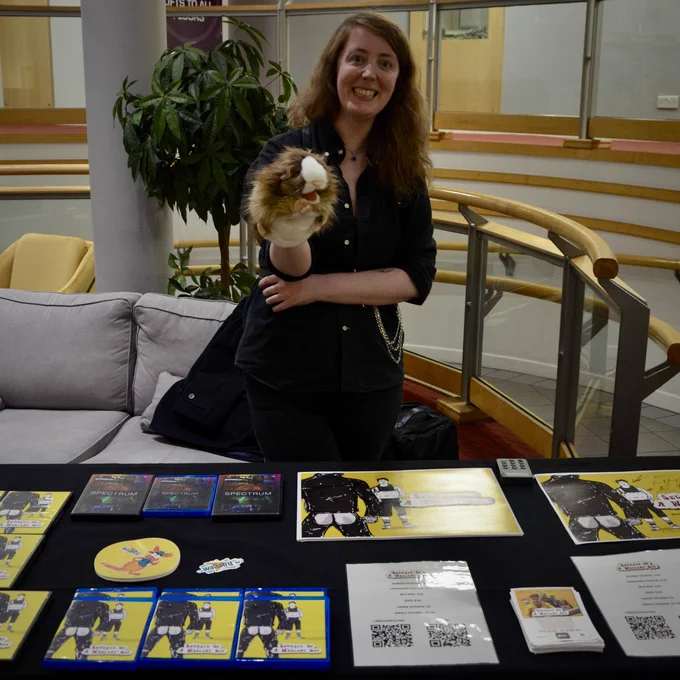 Katie Widdowson with a guinea pig puppet stood behind a table of Secrets of a Wallaby Boy merchandise.