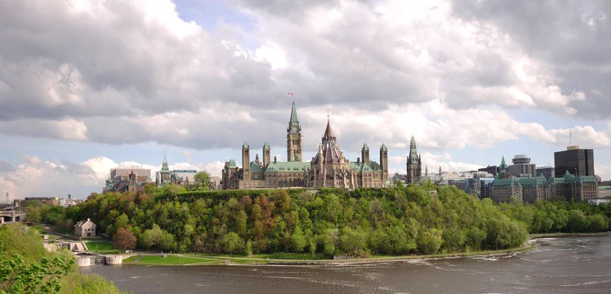 The photo showcases the iconic Centre Block of the Parliament Buildings in Ottawa, Canada, viewed from across the Ottawa River. The building is characterized by its Gothic Revival architecture, featuring multiple towers and spires, with the most prominent being the Peace Tower at the center, adorned with a Canadian flag at its peak. The structure is set against a backdrop of lush greenery and trees, with the river in the foreground, reflecting parts of the scene. The sky above is partly cloudy
