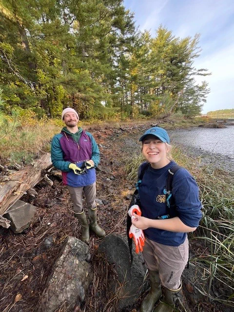 Two people standing together, smiling, on a salt marsh shoreline with water and trees in the background