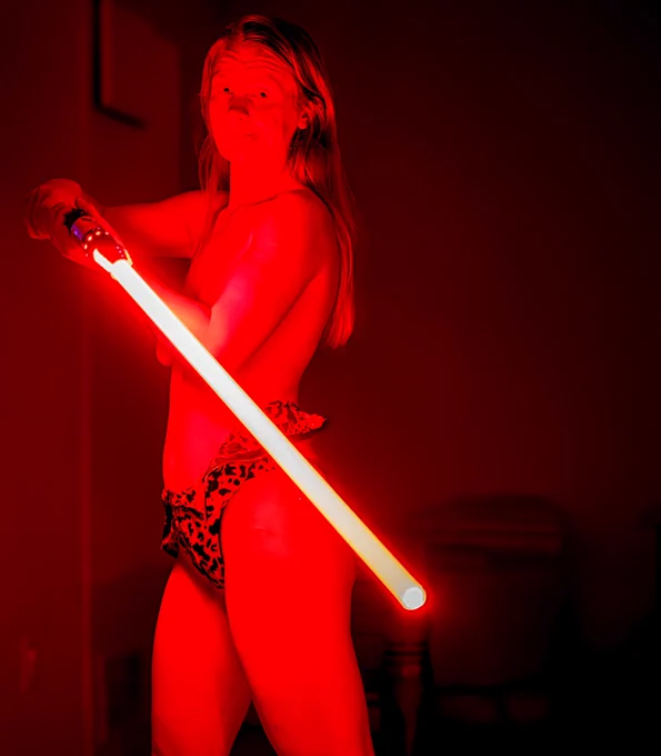 Lily, with her vibrant red ginger hair glowing under the intense red lighting, wields a bright red lightsaber in a dynamic pose. Her freckled skin catches the dramatic glow of the saber, while she wears a leopard-print bikini bottom that complements her playful yet fierce energy. Her body is slightly turned, emphasizing her toned figure against the dark, minimalist background, creating a bold and commanding presence.