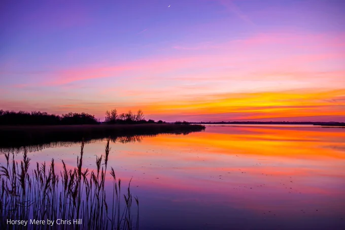 A photograph showing the still waters of Horsey Mere illuminated by vibrant orange, pink and purple colours. Silhouetted reeds and vegetation are visible to the left of the frame.