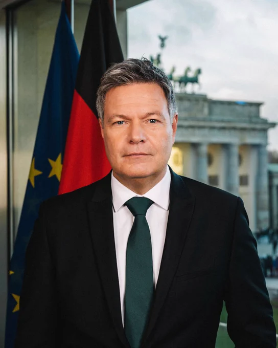 Ein Foto von Robert Habeck. Hinter ihm zu sehen sind eine Deutschland- und eine Europaflagge. Außerdem das Brandenburger Tor. Er schaut direkt in die Kamera.