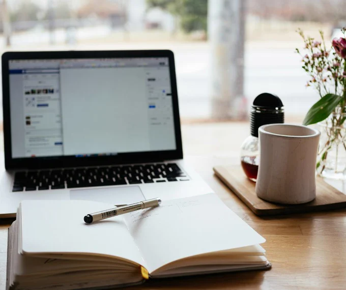 A laptop on a wooden desk with a notepad open in front of it. There is a coffee mug beside it ready to go!