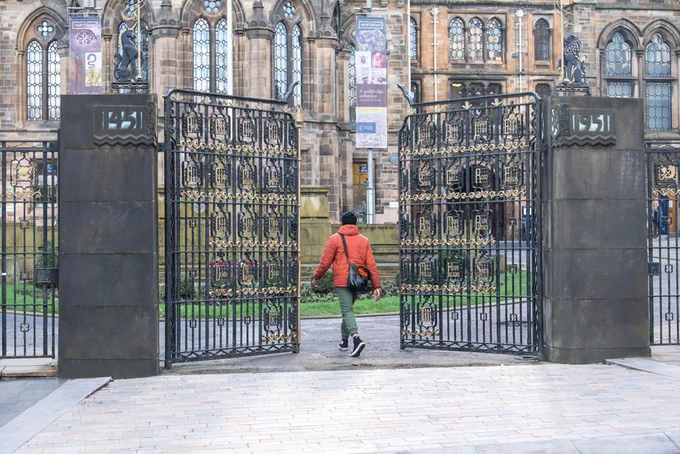 A person in a red winter coat and green trousers, walking through two large iron gates which lead towards the University of Glasgow Gilbert Scott building.