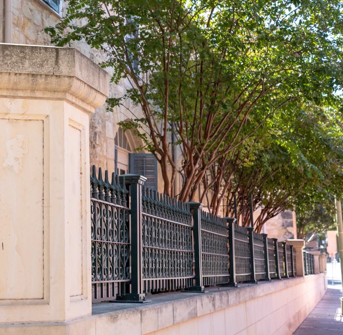 Elegant black wrought iron fence along a sunlit sidewalk beside a historic stone building with a leafy tree in the background.