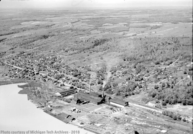 Aerial view of a smelter with a house- and tree-lined hillside behind it