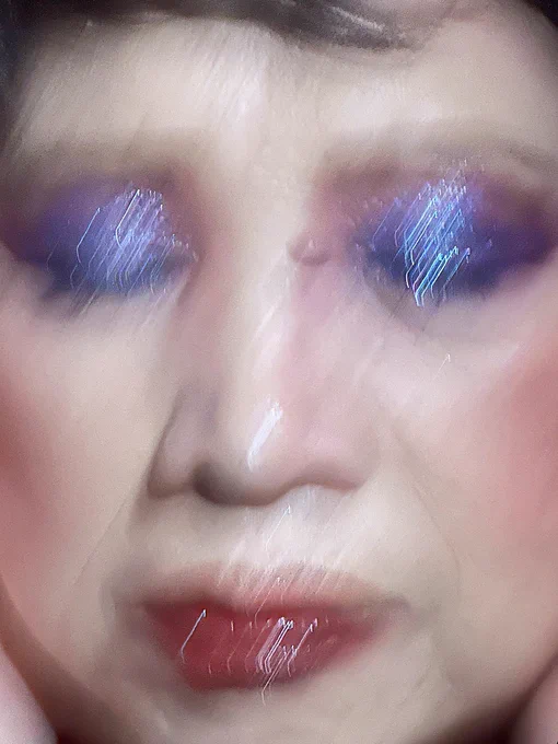A blurry close-up portrait of a Chinese grandmother with eyes shut. She wears glittery purple eye shadow and shimmery red lip gloss.