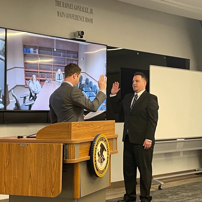U.S. Attorney Josh Hurwit swearing in a new HIDTA SAUSA pictured behind a podium with a DOJ seal. A screen in the background shows a conference room with 2 people at the table.