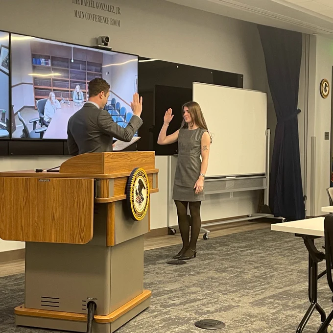 U.S. Attorney Josh Hurwit swearing in a new legal administration specialist pictured behind a podium with a DOJ seal. A screen in the background shows a conference room with 2 people at the table.