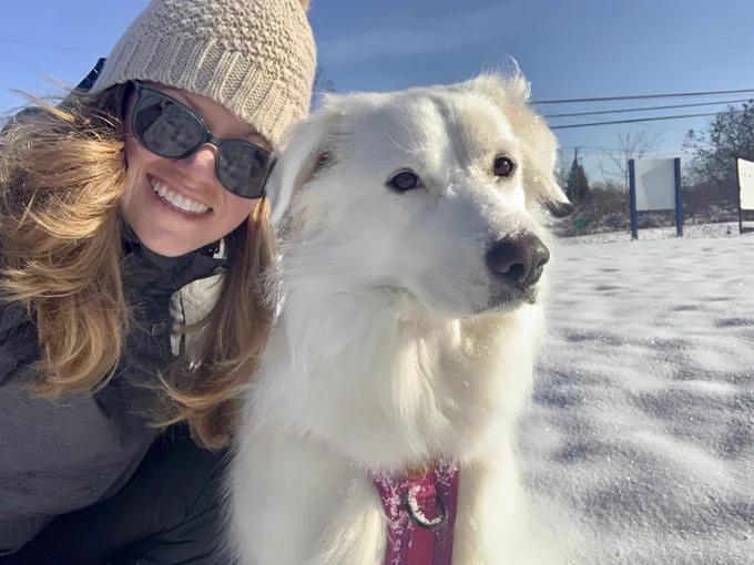 Sarah smiles while posing for a selfie with a white dog in a pink harness. She is wearing a cream-colored knit beanie and sunglasses. Credit: Sarah Peacock