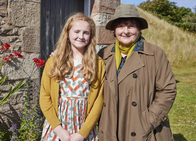 Two individuals smiling and standing outdoors next to a stone building with red flowers and greenery in the background. The person on the left, a young girl with long hair, wears a floral dress and yellow cardigan. The person on the right, Brenda Blethyn, is dressed in a brown coat and green hat.