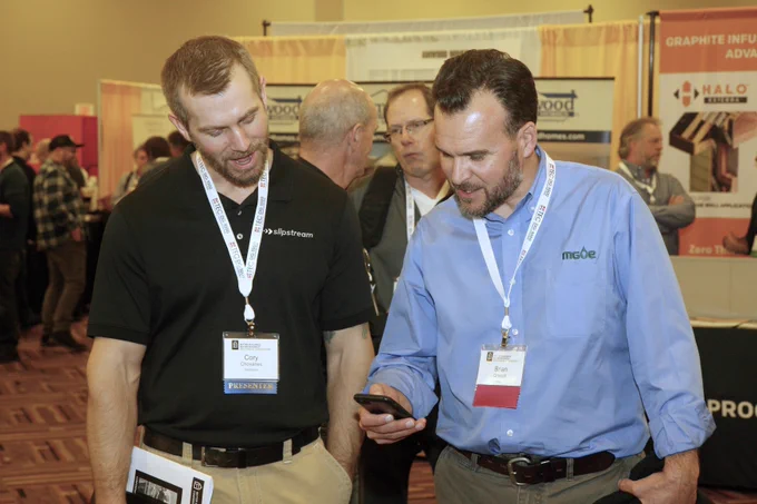 Two individuals, one holding a smartphone, engage in a conversation at a business expo with booths and other attendees visible in the background. 