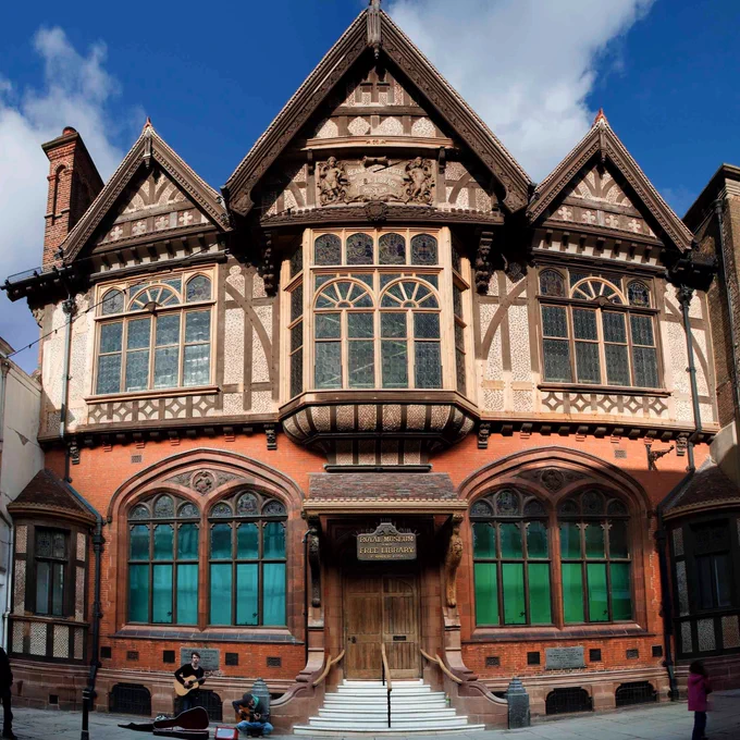 The Beaney, a Tudor style building with red brickwork, decorative woodwork, arched windows and a central flight of steps to a roofed entrance. The roof has three gables the largest in the middle.