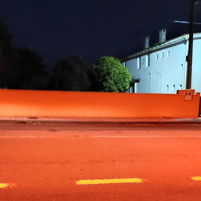 En la noche, carretera de color rojo con líneas amarillas. Detrás hay una casa grande iluminada de color blanco con un árbol verde. Más árboles oscuros y un cielo negro.