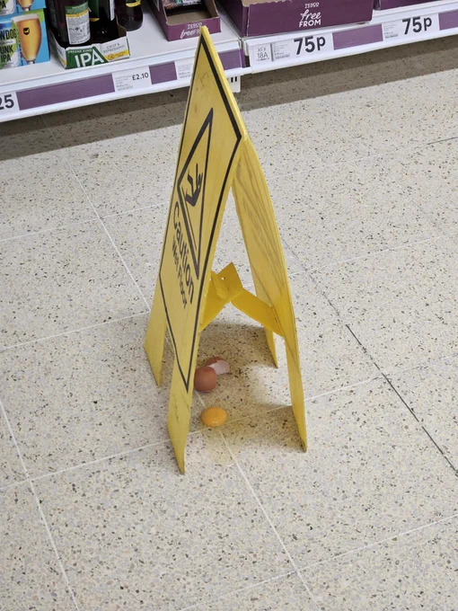 A big yellow safety sign warning for a wet floor, covering a single broken egg on what appears to be the floor of a supermarket.