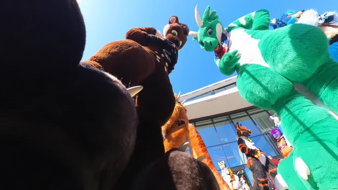 A big, cream-and-brown colored Otter Fursuiter named Blackymoon is standing next to a green-and-white Dragon Fursuiter namend DC. The view onto them both is shown from a very low-angle point of view with one of Blackymoons footpaws covering a third of the whole photo. Both Blackymoon and DC are looking down at the camera, making the whole photo feel as if they are two giants. Behind them are several other Fursuiters walking around.