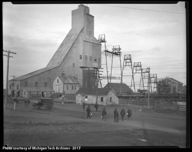 Men walking past a vehicle with lunch pails as a mine shafthouse stands in the background