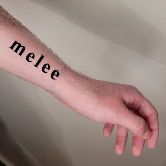 photo of a fresh tattoo on the wrist of a person. the text reads, "melee" in times new roman bold font. it is centred on the inside wrist in line with the thumb.