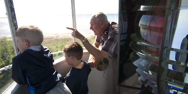 A lighthouse keeper points out from the lantern room of a lighthouse, showing two small children the view.