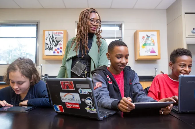 A group of three students sitting at a table use laptops and tablets as their teacher stands looking on from behind them. 
