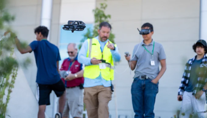 An adult and student using VR drone equipment in the background while a drone hovers in focus in the foreground. 
