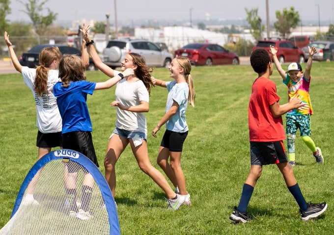 A group of kids celebrating and high diving on in a field with a soccer net in the fore ground. 
