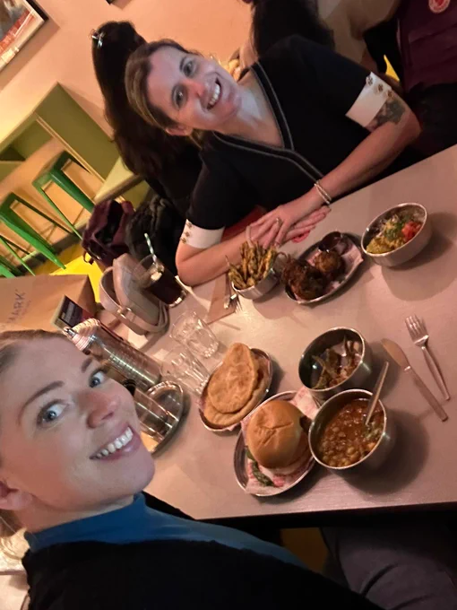 Two women smile at a camera in a selfie image. They are in a restaurant called Bundobust. There are 7 small plates of Indian street food on the table. 