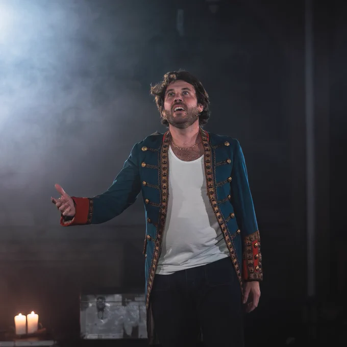 An individual passionately performing on stage, dressed in a decorative jacket and white shirt, with candles and a dimly lit background setting.