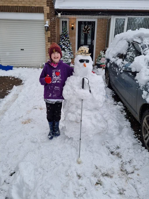 Eli, a 9-year-old boy, wearing a hoody, hat, and gloves, stands next to a large snowman that is dressed in a straw hat, sunglasses, and holding a cane.