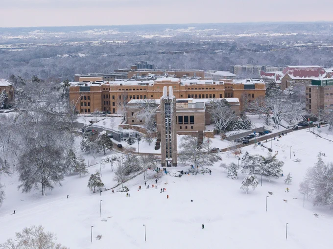Aerial view of the snow-covered Lawrence campus. The campanile is the central focus including some individuals sledding down the Hill. 