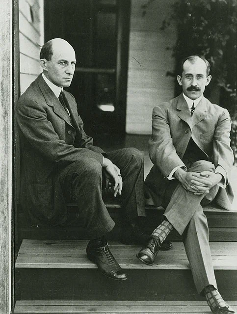 Black and white photograph of two men sitting on the steps of a house, dressed in early 20th-century attire.