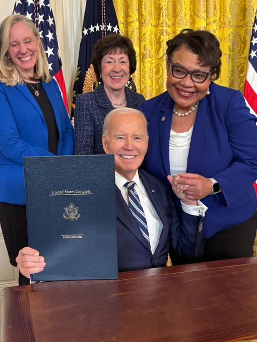 President Joe Biden smiles surrounded by Rep. Abigail Spanberger, Sen Susan Collins, and NEA Vice President Princess Moss. He holds a document titled "United States Congress." They are in a room with ornate yellow curtains and American flags in the background.