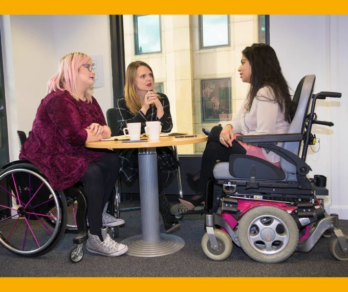 Three people are sitting around a desk in an office. Two of the people are wheelchair users. They are trainers for Enhance the UK
