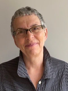 Photo of Nicole, smiling. She has short grey hair and glasses, and wears a dark blue shirt.