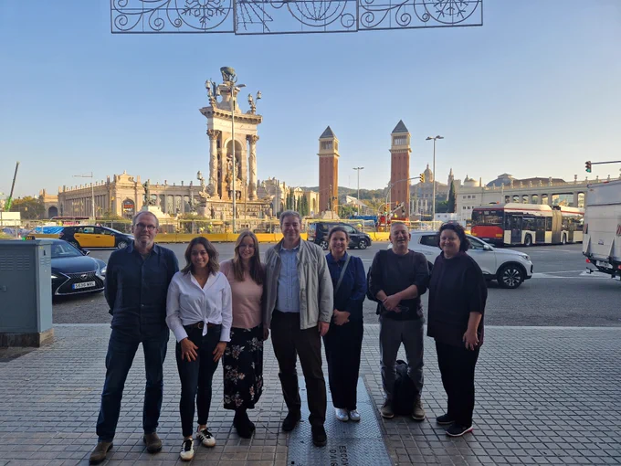 7 members of the EuroQol comms team pose in on the side of a busy road with cars driving. Behind them is the Place d’Espanya in Barcelona, Spain, underneath a clear blue sky.