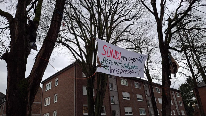 In zwei Bäumen sitzen Menschen, eine Person in einer Art Hängematte, die andere zwischen zwei Ästen. Im Hintergrund ist ein weiterer Baum zu sehen. \nZwischen den Bäumen ist ein Transparent gespannt mit Aufschrift "SÜNDIgen gegen RWE. Besetzen, sabotieren, freisprechen!" und einem gemalten Ast. Der Himmel ist grau und Wohngebäude sind im HIntergrund auch zu sehen.