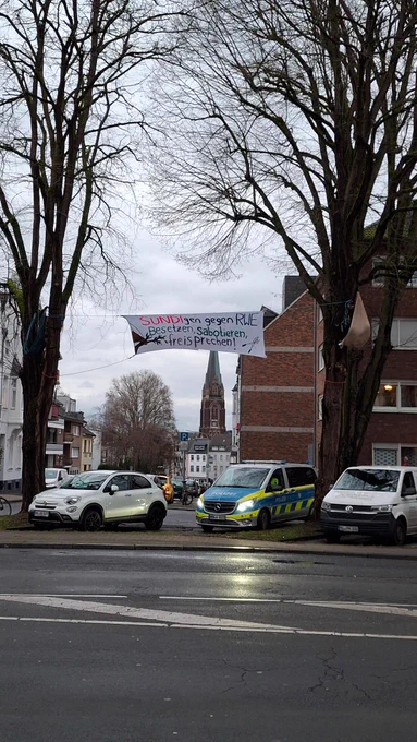 Zwei Bäume ragen hoch in den Himmel, darunter sind drei Autos, das mittlere eine Wanne von der Polizei. \nZwischen den Bäumen ist ein Transparent gespannt mit Aufschrift "SÜNDIgen gegen RWE. Besetzen, sabotieren, freisprechen!" und einem gemalten Ast. Der Himmel ist grau, die Straße im Vordergrund auch. Im Hintergrund sind Gebäude zu sehen.