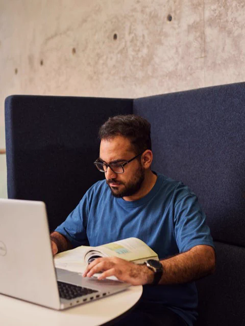 Person in a blue shirt studying from a book and using a laptop in a modern workspace.