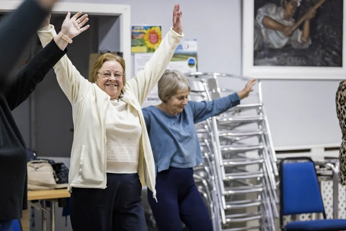 Two older women participate in a Dance On session in a community venue. The woman on the left raises her arms joyfully, wearing a light jumper and glasses, while the woman on the right looks on, smiling, and is dressed in a blue top. Metal folding chairs are stacked in the background.