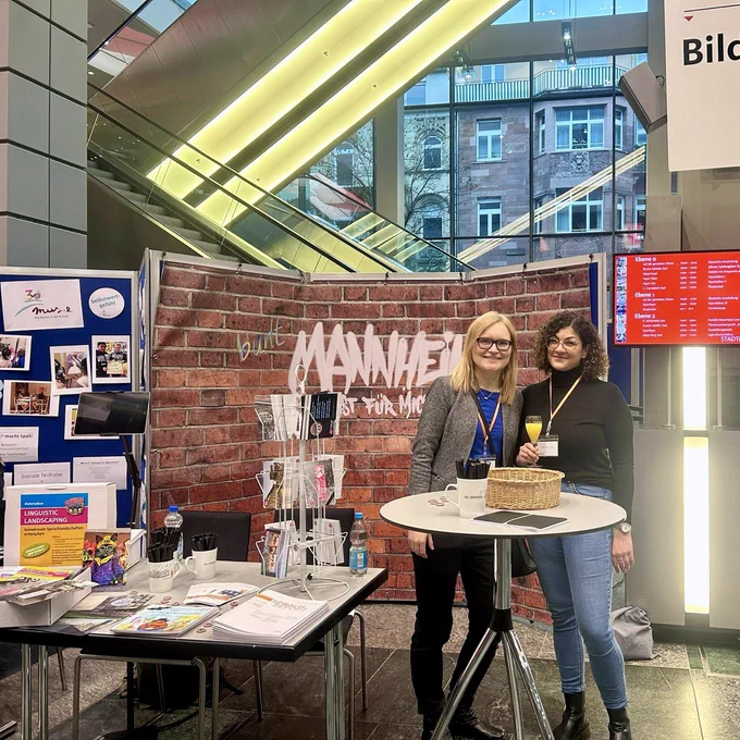Man sieht den Stand des IDS beim Neujahrsempfang der Stadt Mannheim 2025, im Vordergrund einen Tisch mit der Materialbox für Schulen sowie mit Flyern, Ausgaben der Zeitschrift Sprachreport und des Sprach-Checker-Kinderbuchs und einem Postkartenständer. Rechts daneben stehen an einem Stehtisch die Mitarbeiterinnen Janin Rössel und Angelina Steber. Im Hintergrund sieht man eine auf einer Stellwand aufgespannte Plane in Backsteinoptik mit dem Schriftzug "Mannheim ist für mich…", die von den Besucherinnen und Besuchern als Mannheimer Sprachlandschaft gestaltet werden kann. Rolltreppen sind sichtbar hinter dem Stand und angrenzend der nächste Stand.