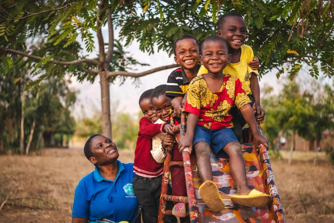 Five children smiling and playing on a playground slide with an adult supervising from the side, in a rural setting.