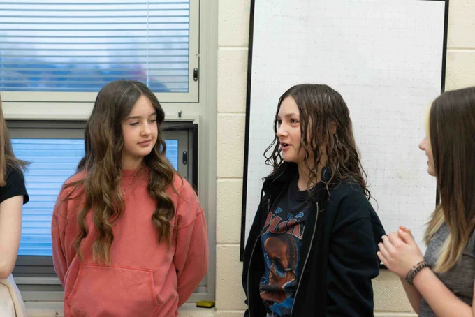 Middle school students standing in a classroom. One is speaking, another is looking at them and listening.