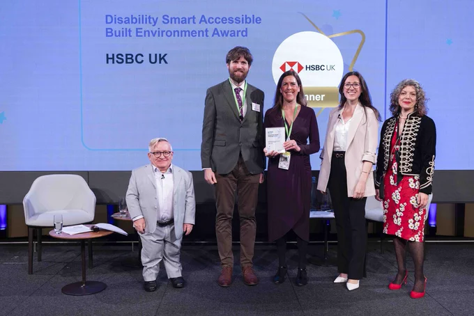 Five people posing on stage at an event. The person in the middle is holding an award. Behind the group is a screen with the HSBC UK logo and the words: 'Disability Smart Accessible Built Environment Award: HSBC UK.'