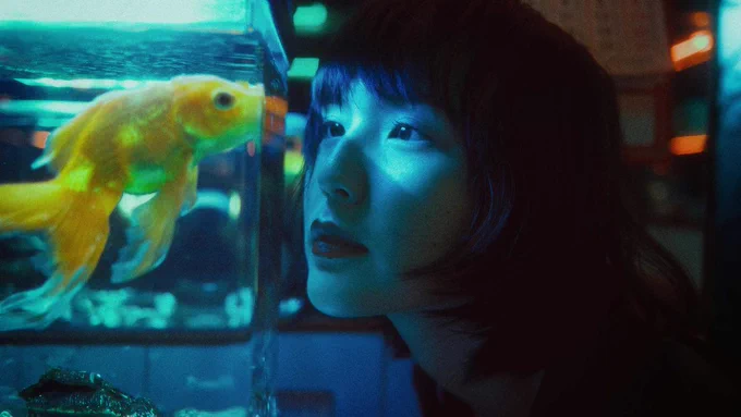A goldfish puts it's mouth on the edge of it's tank, a asian woman looks at it eye to eye. it's a dark blue light, the goldfish is bright.
